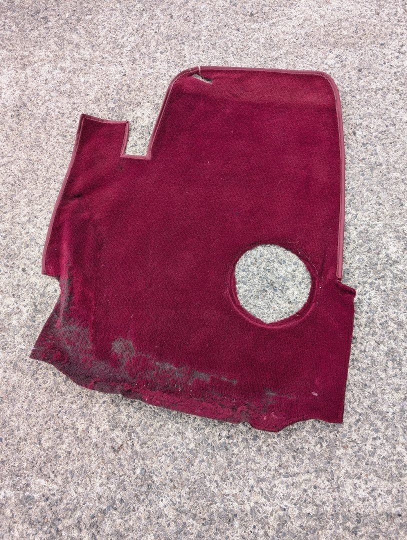One panel of the original red carpet, lifted out whole — the bound edge, the gear lever aperture and the cut-outs all intact, ready to be laid on the new roll and cut round.